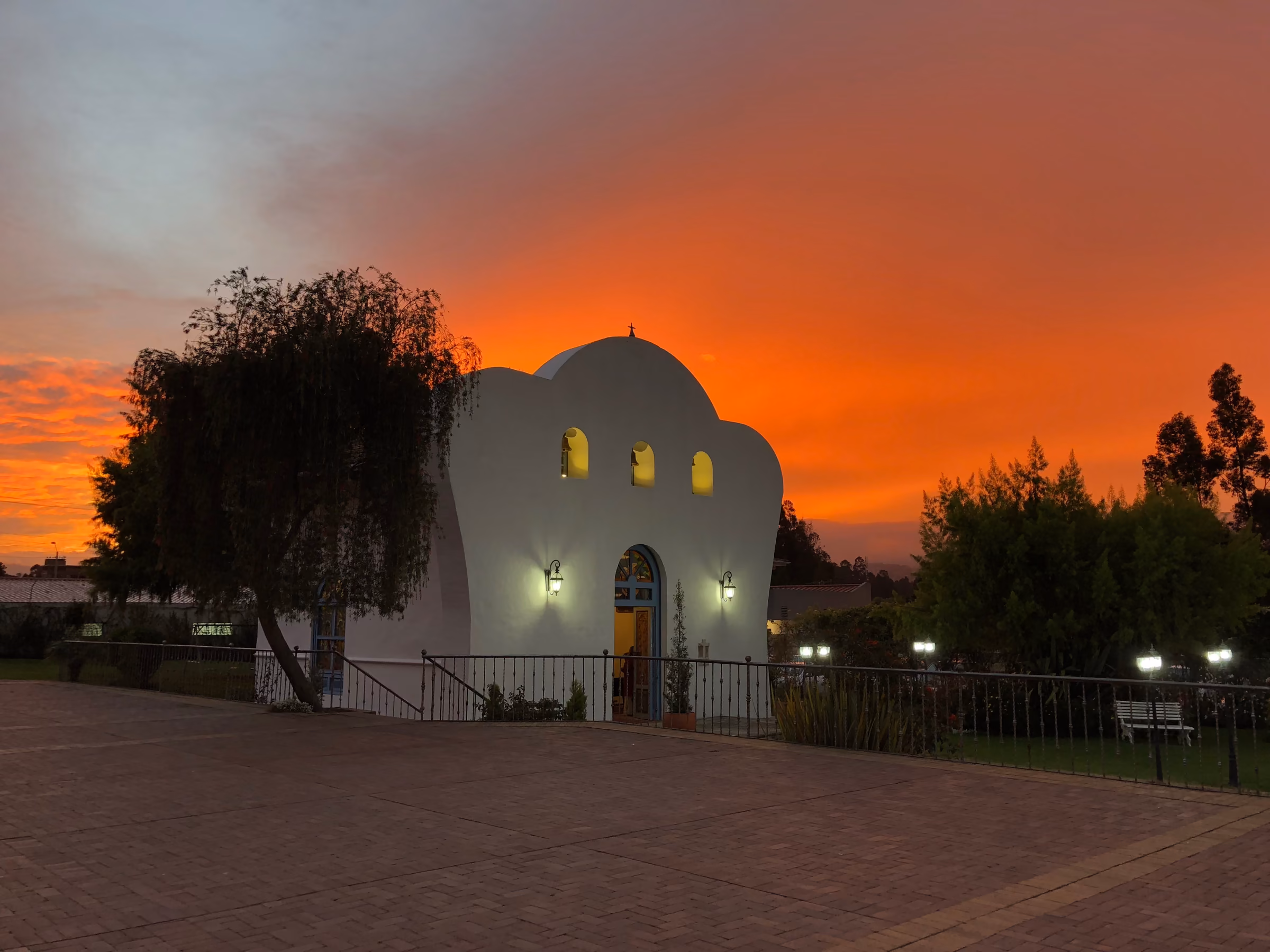 The chapel lit beneath a blazing orange sky, with the willow and the brick plaza in the foreground.