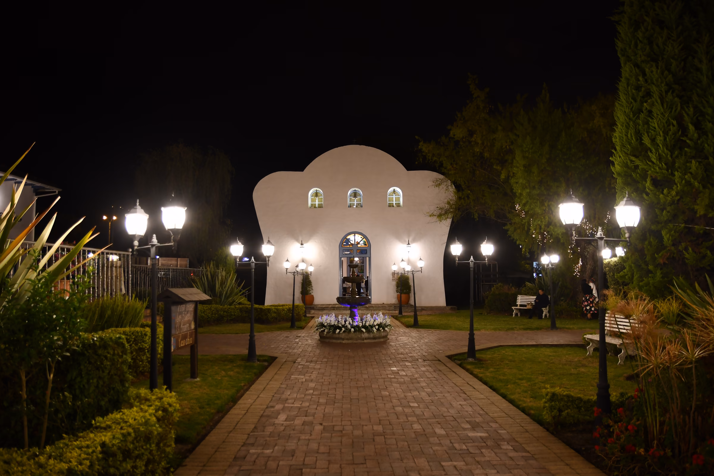 The chapel lit at night at the end of the brick walkway, lined with lamp posts and planters.