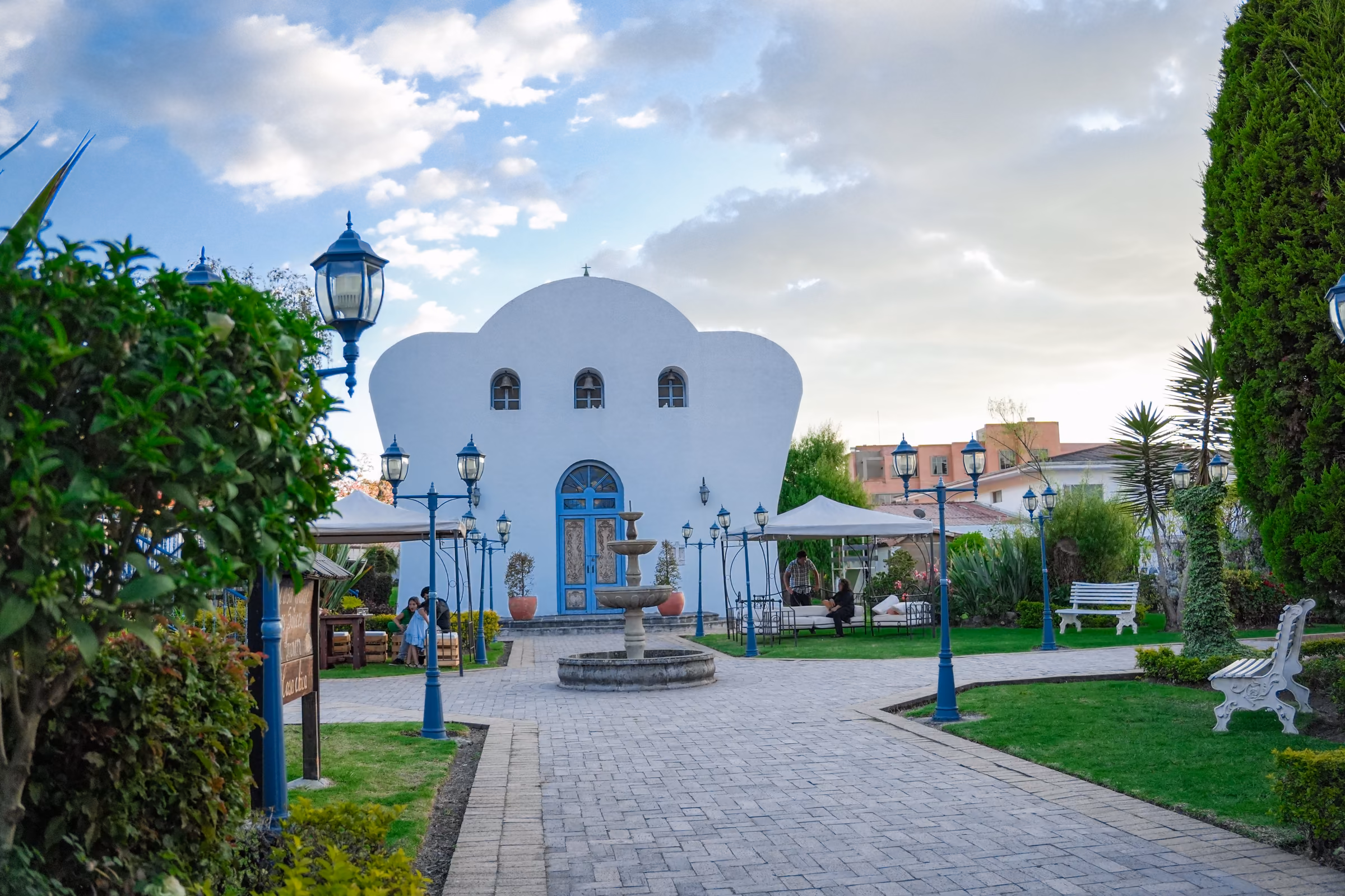 The white chapel at the end of the garden, with its stone fountain, blue lamp posts and benches under a cloudy sky.