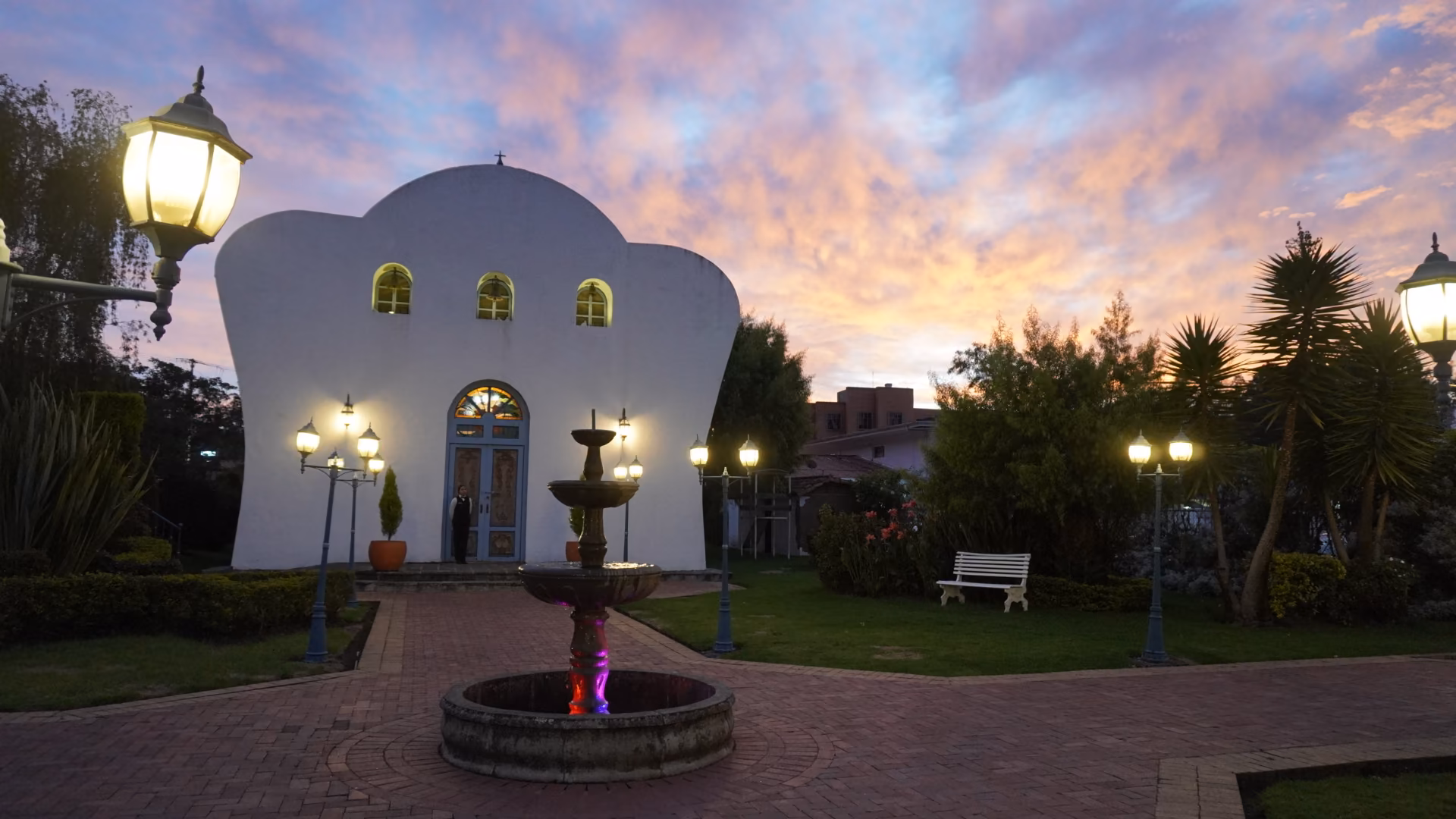 The fountain and chapel lit beneath a pink sunset sky, with lamp posts glowing across the garden.