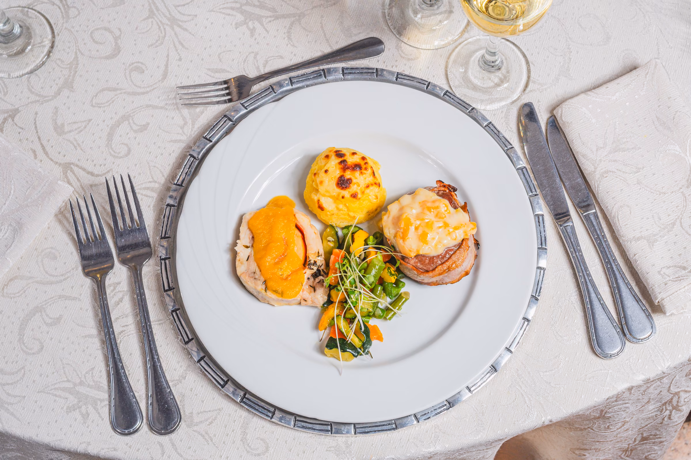 Plato servido con pollo relleno en salsa de zanahoria, medallón envuelto en tocino, timbal de papa gratinado y salteado de vegetales.