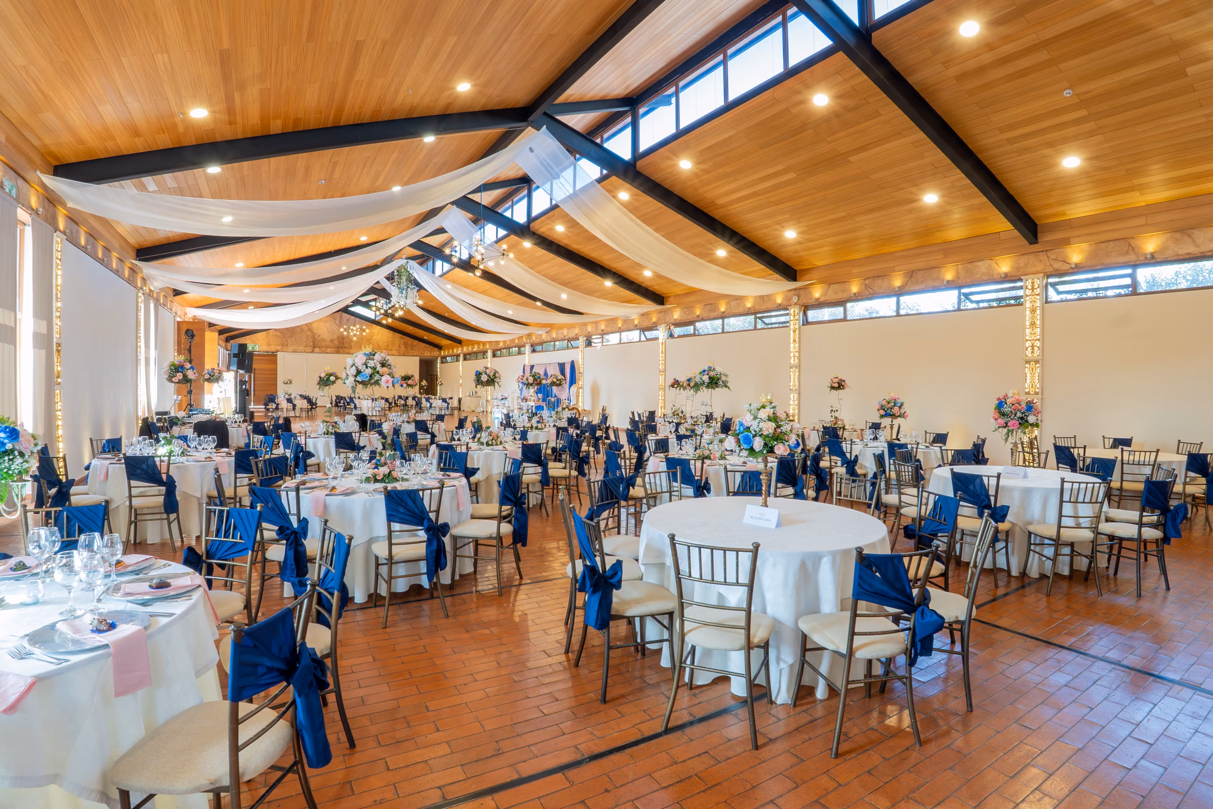 Round tables dressed with navy sashes and pastel flowers under the pale timber ceiling with black beams, lit by daylight.