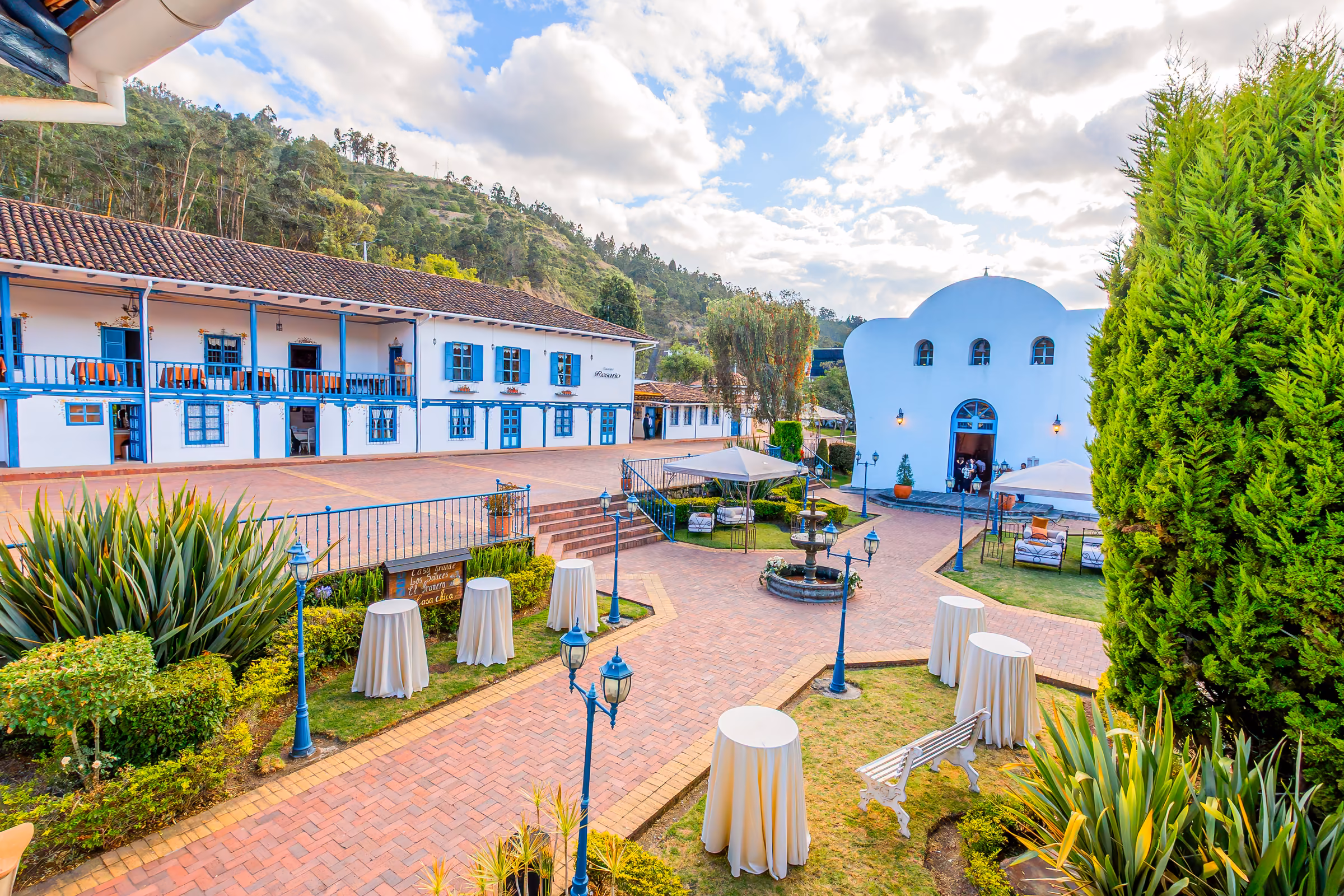 A brick plaza with a fountain and white-draped cocktail tables set between the blue-balconied main house and the whitewashed chapel.